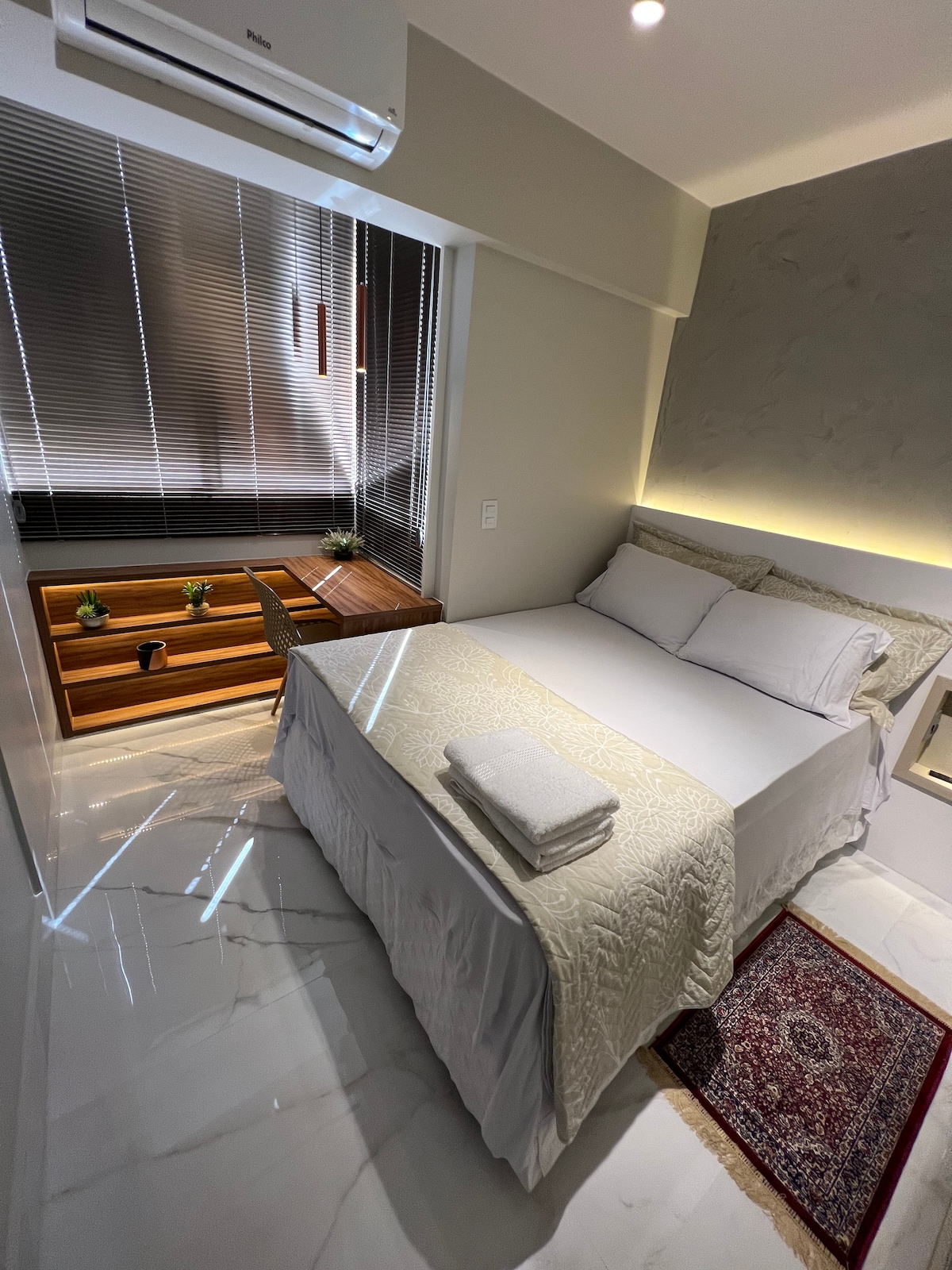A well-organized bedroom is presented, featuring a spacious bed with white linens, a decorative throw, and neatly arranged towels. Natural light filters through window blinds, illuminating the room. A wooden desk and small indoor plants add a touch of warmth to the modern decor.