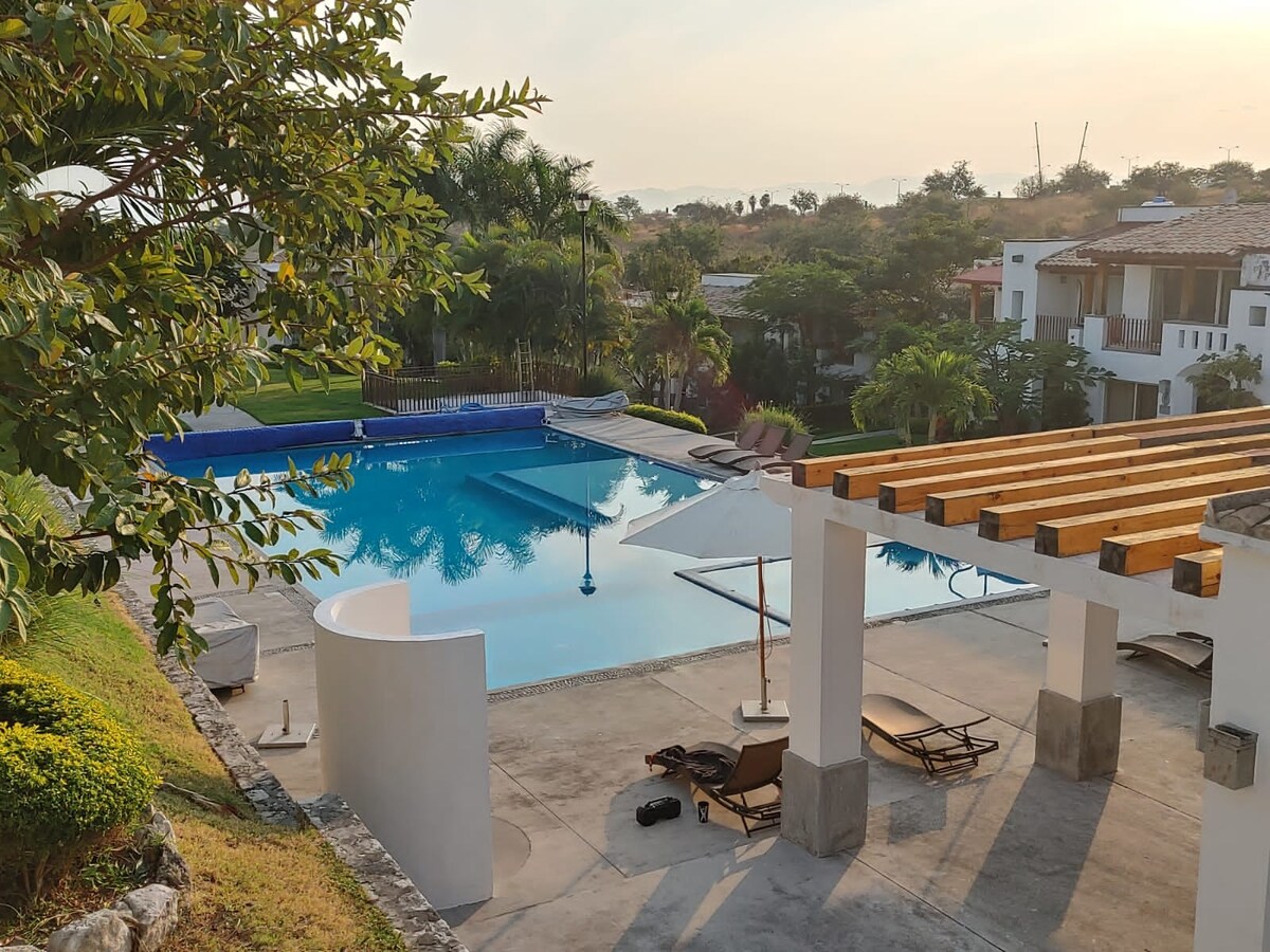A spacious outdoor area features a large swimming pool surrounded by lush greenery. Sun loungers are placed near the pool area, and an outdoor structure with wooden beams provides shade. Soft lighting enhances the serene atmosphere as the sun sets over the horizon.
