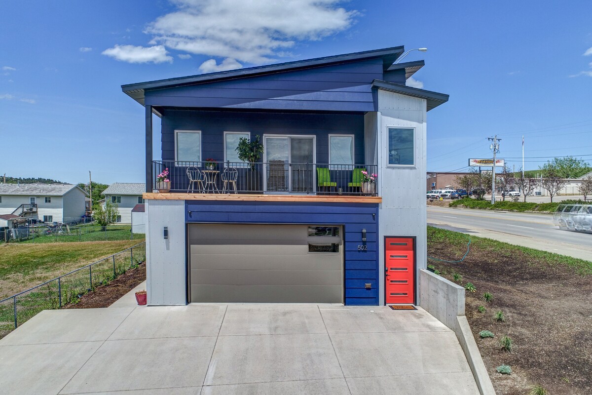 A modern house is presented from an elevated angle, showcasing a two-tone exterior in blue and gray. A spacious front deck features green chairs around a table, providing a setting for outdoor relaxation. A red door adds a vibrant accent to the entrance.