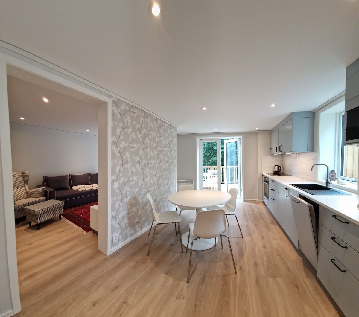 A modern kitchen and dining area is presented, featuring light cabinetry and a round white dining table with four chairs. Sunlight enters through sliding glass doors, leading to a private balcony. A cozy living area with a sofa is visible in the background.