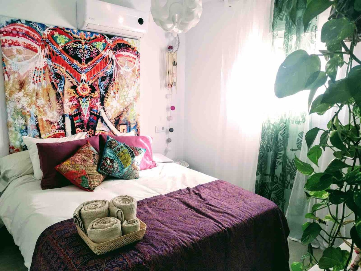 A bright bedroom features a spacious bed dressed with layered linens, including a purple throw and decorative pillows. A vibrant elephant tapestry adorns the wall, while sunlight filters through sheer curtains. Soft plants add a touch of nature to the cozy space.