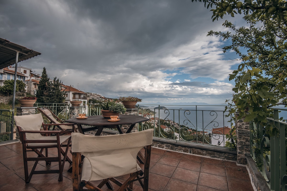 A spacious balcony is presented, featuring a round table set with plates and cups. Four light-colored chairs surround the table, and potted plants add a touch of greenery. In the background, the Aegean Sea and distant landforms are visible under a partly cloudy sky.