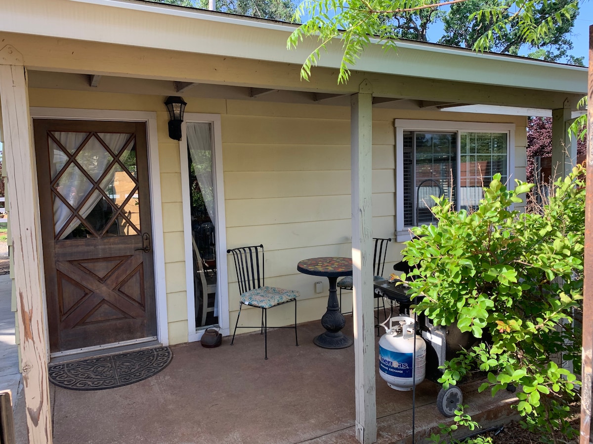 A covered porch features a wooden front door and a small dining table with two chairs. Plants flank the entrance, adding greenery, while a gas grill is positioned nearby. Windows allow natural light into the space, enhancing the welcoming appearance.