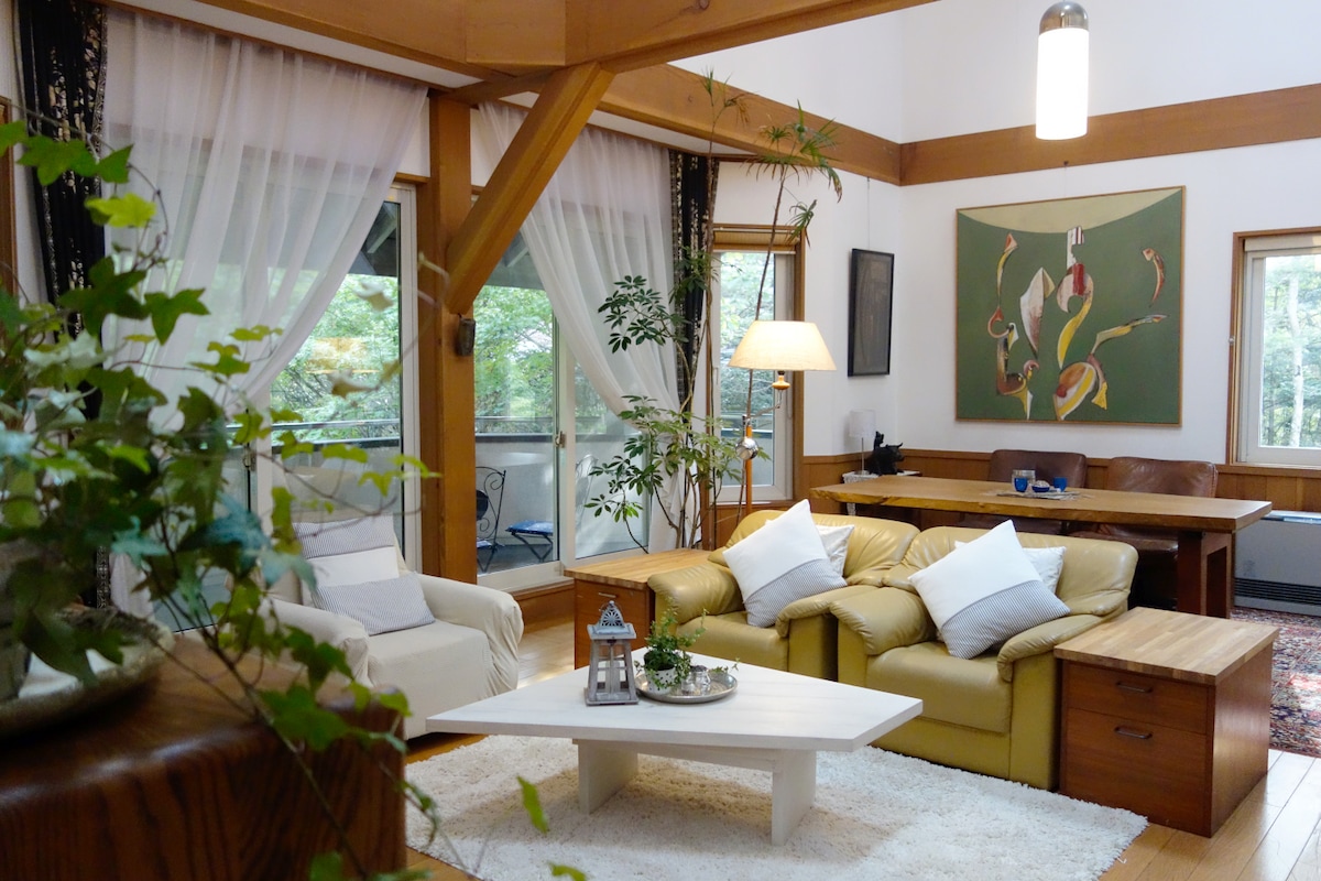 A spacious living area features large windows allowing natural light to fill the room. A white area rug is positioned beneath a low coffee table, surrounded by a yellow sofa and armchairs. Potted plants add greenery, and a warm table lamp provides soft illumination.