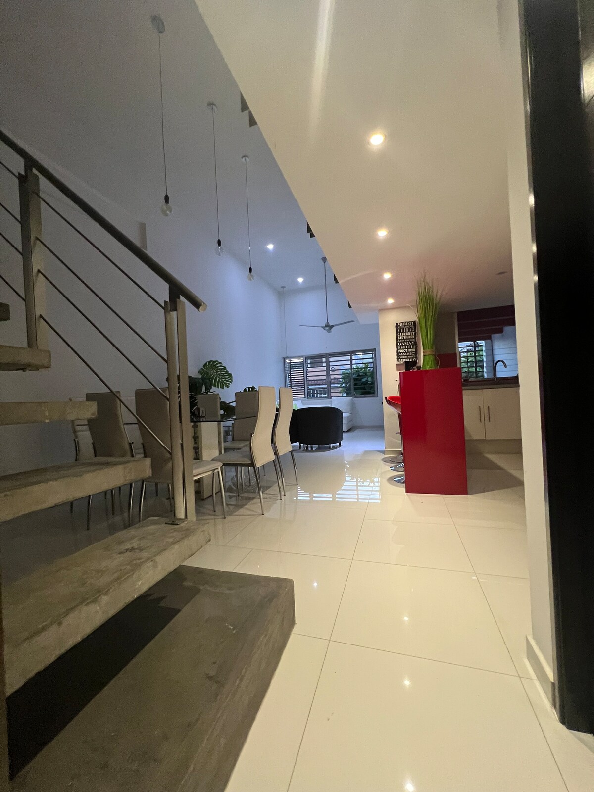 A modern dining area is visible, featuring a long table surrounded by several chairs. Bright lighting enhances the spaciousness of the room, while a red kitchen island is pictured in the background. Large windows provide natural light, complemented by minimalistic decor.