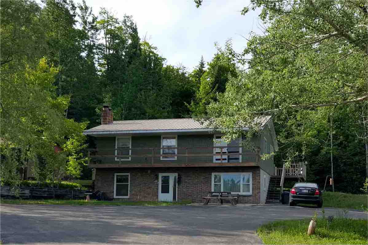 A two-story lodge is nestled among trees, featuring a gently sloping yard. The exterior showcases a combination of wood and brick, with a shaded porch and large windows. A gravel driveway leads to a parked vehicle, and outdoor seating is visible in front.