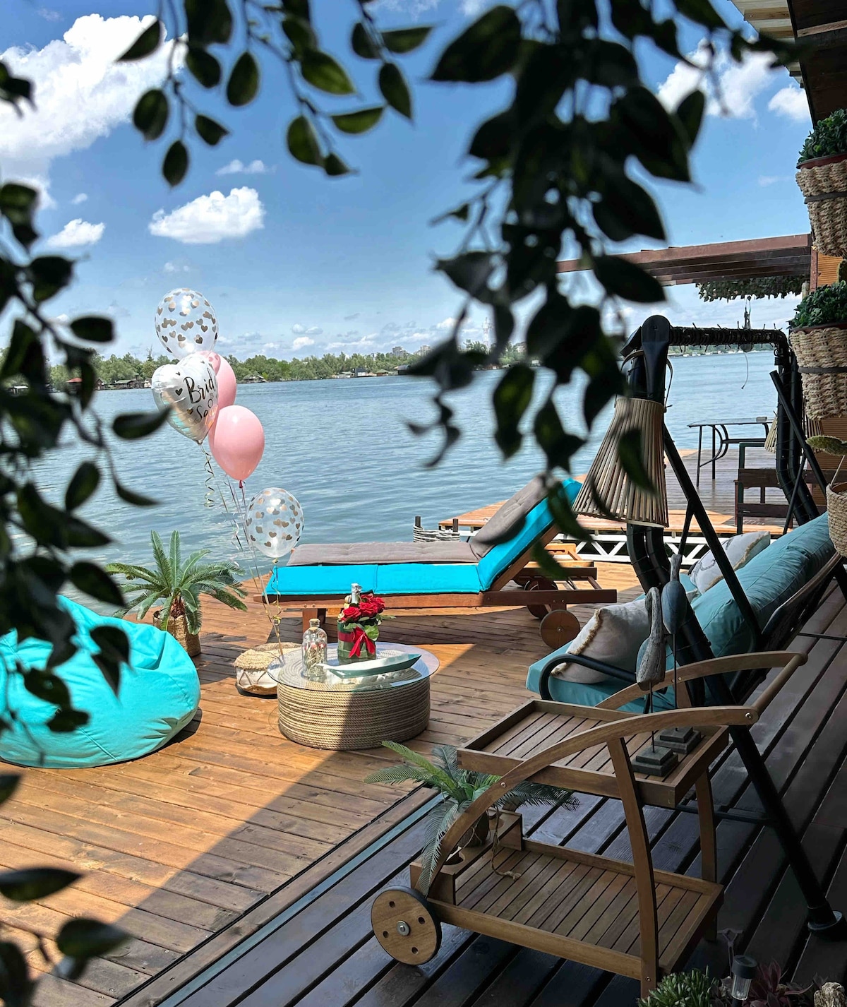 A wooden deck extends over the water, featuring comfortable lounge chairs and colorful bean bags. A small table is centrally positioned, adorned with fresh flowers. Balloons in soft colors float nearby, while greenery frames the serene view of the river.