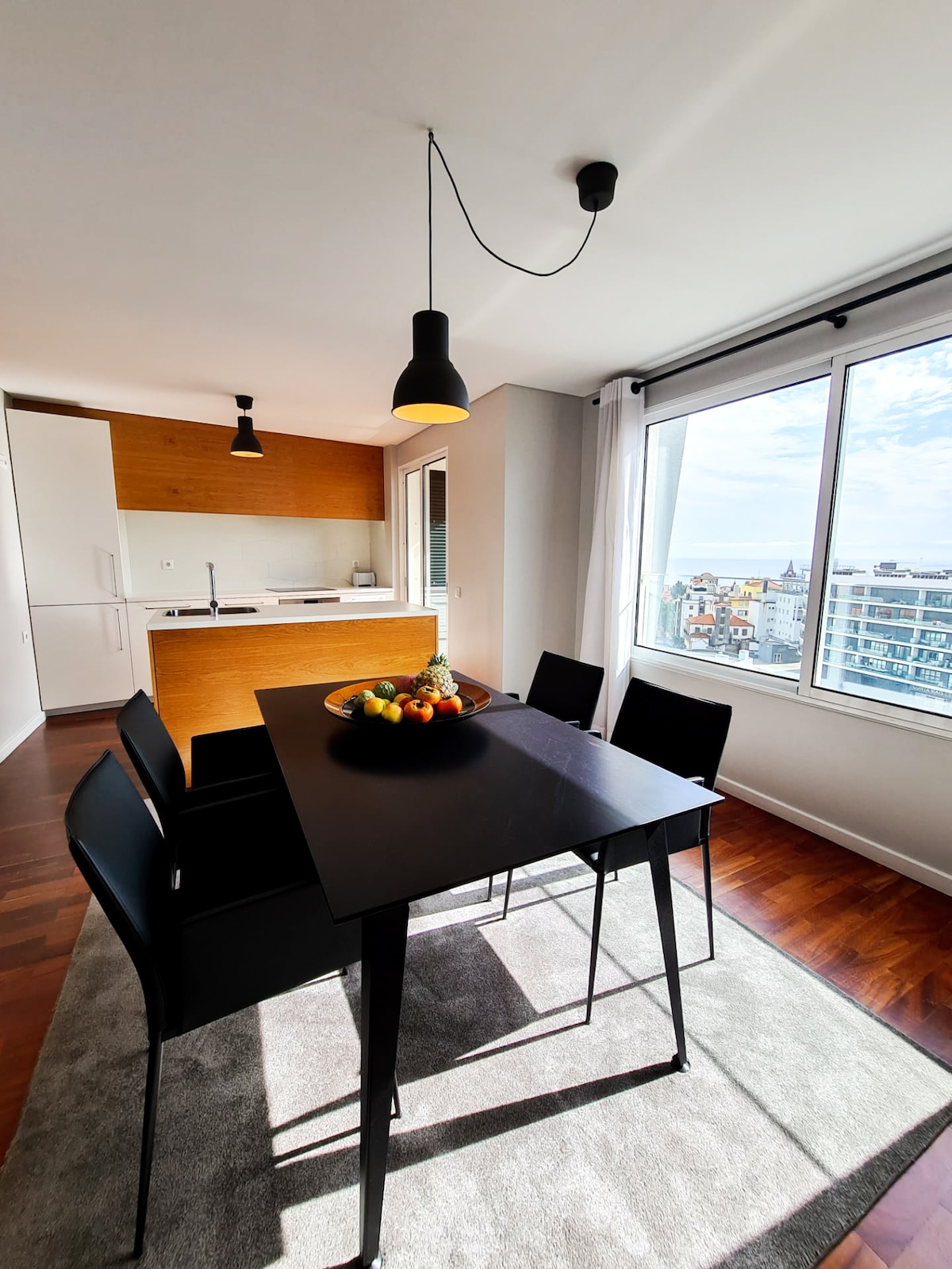 A dining area is presented with a sleek black table surrounded by four chairs. A bowl of colorful fruit is centrally placed on the table. Natural light fills the space through a large window, offering views of the city and brightening the modern kitchen in the background.