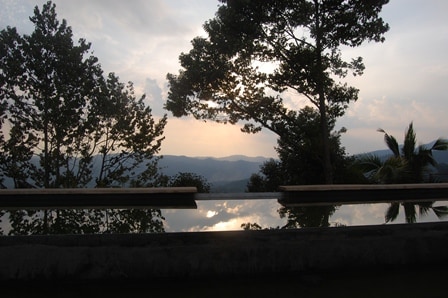 The serene scene features a tranquil swimming pool reflecting the soft hues of a sunset, framed by lush trees. The distant hills create a peaceful backdrop, enhancing the sense of calm offered by this natural setting.