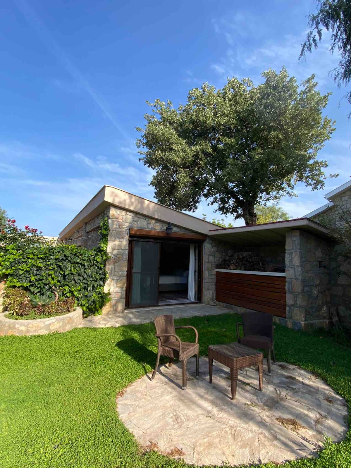 A stone cottage is framed by lush greenery, featuring a small seating area with two chairs positioned on a circular stone patio. Nearby, a large tree casts shade over the space, inviting relaxation and connection with nature.