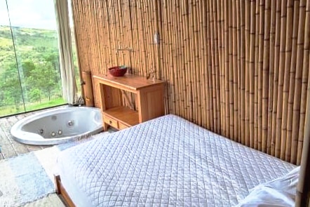 A rustic sleeping area is framed by bamboo walls, featuring a spacious bed with a light quilt. A wooden shelf is positioned beside the bed, holding a decorative bowl. A jetted tub is situated nearby, offering views of the surrounding greenery through large glass panels.