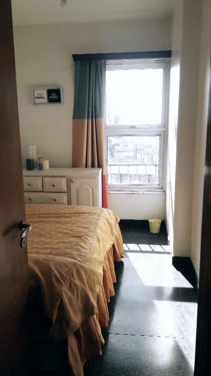 A cozy bedroom is shown, featuring a spacious bed covered with a soft yellow bedspread. Natural light filters through a window adorned with colorful curtains, illuminating a white dresser positioned nearby. A small decorative plant sits in the corner, enhancing the room's atmosphere.