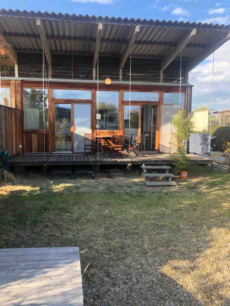 A wooden deck with outdoor seating is visible, surrounded by greenery. Large glass doors allow for natural light to enter the living space, and a covered overhang provides shade. The area is designed for comfort in a serene outdoor environment.
