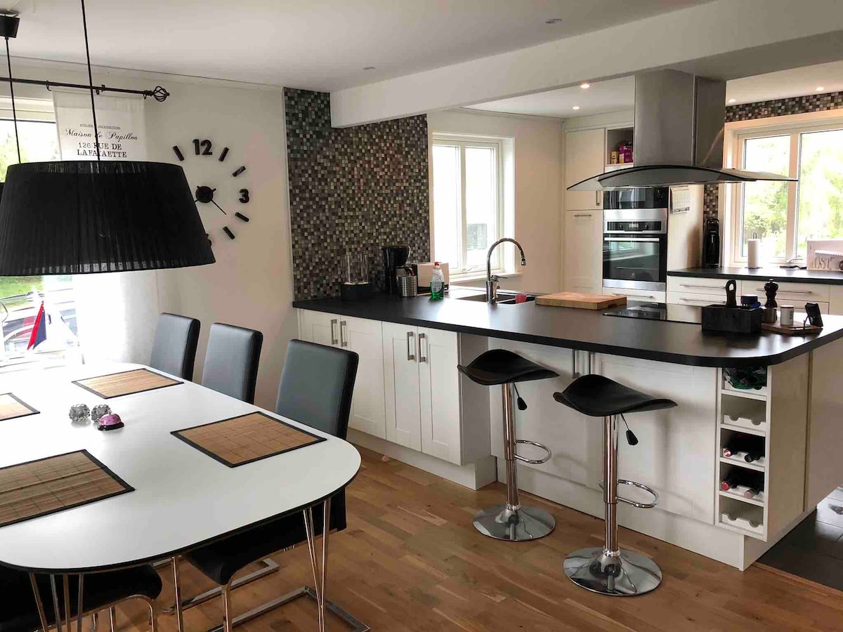 A modern kitchen features a large white dining table with black chairs and a black countertop with bar stools. Natural light enters through multiple windows, highlighting the sleek cabinetry and appliances, including a stove and refrigerator. A decorative clock and mosaic backsplash add visual interest.