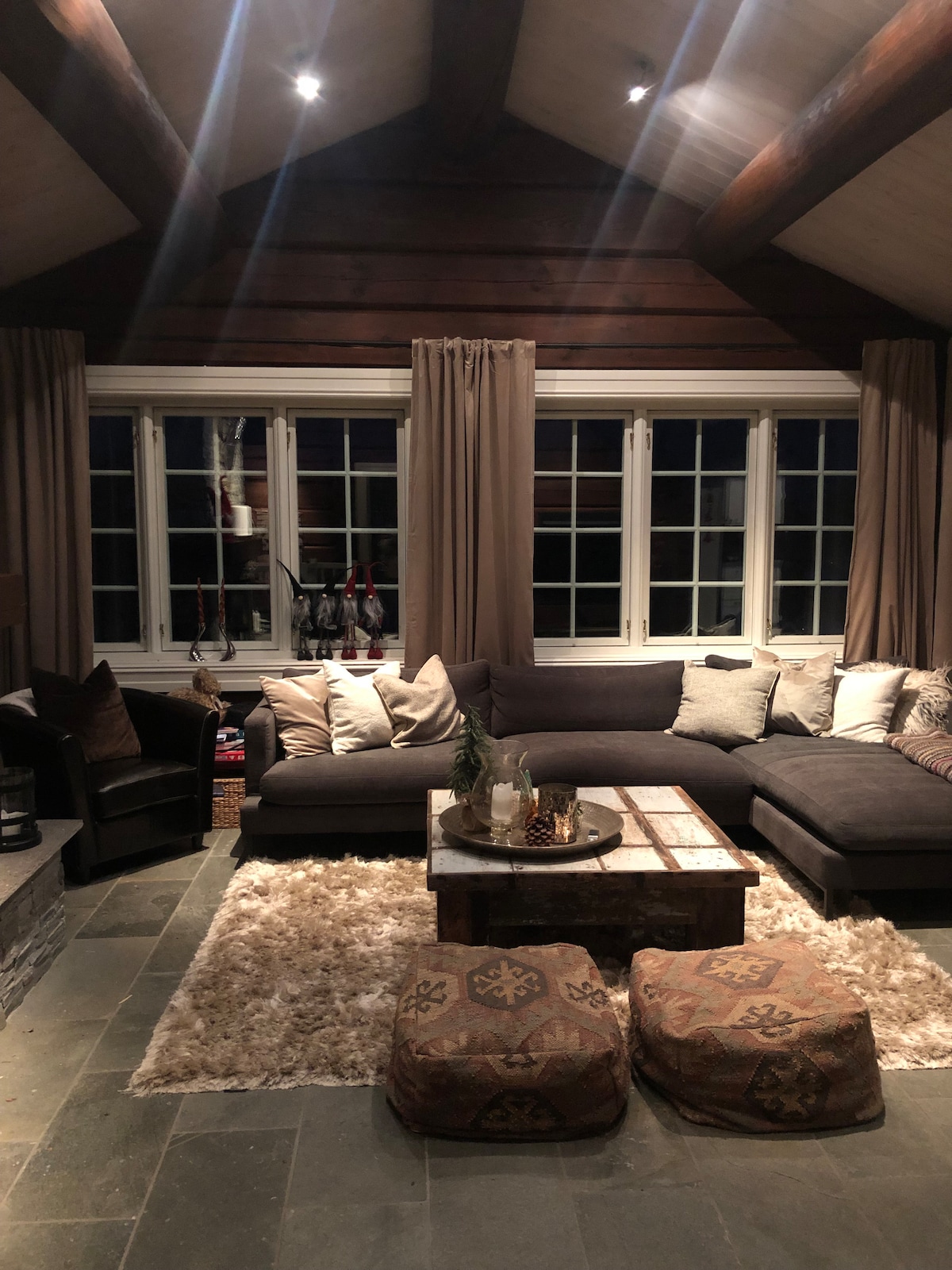 A warmly lit living area features a large, dark sectional sofa adorned with decorative throw pillows. A rustic wooden coffee table sits at the center, complemented by two patterned poufs. Large windows framed with curtains allow for natural light, enhancing the cozy ambiance.
