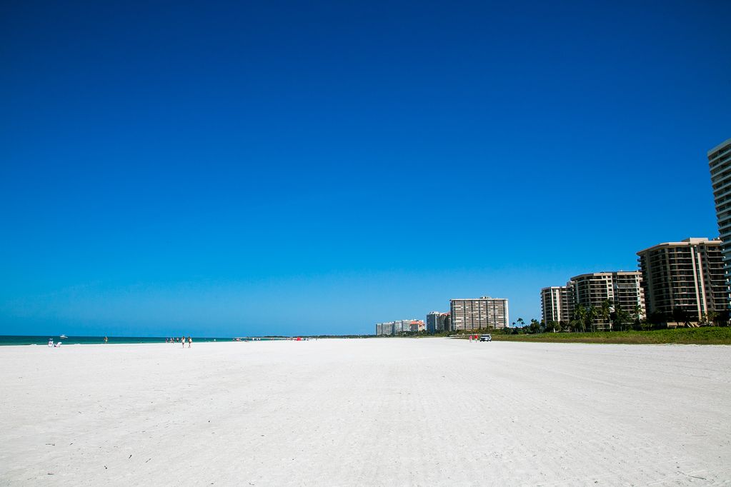 A wide expanse of white sand beach stretches towards the coastline, meeting the clear blue water of the Gulf of Mexico. Tall beachfront buildings with multiple stories stand in the background against a bright blue sky, offering a serene and spacious environment for relaxation.