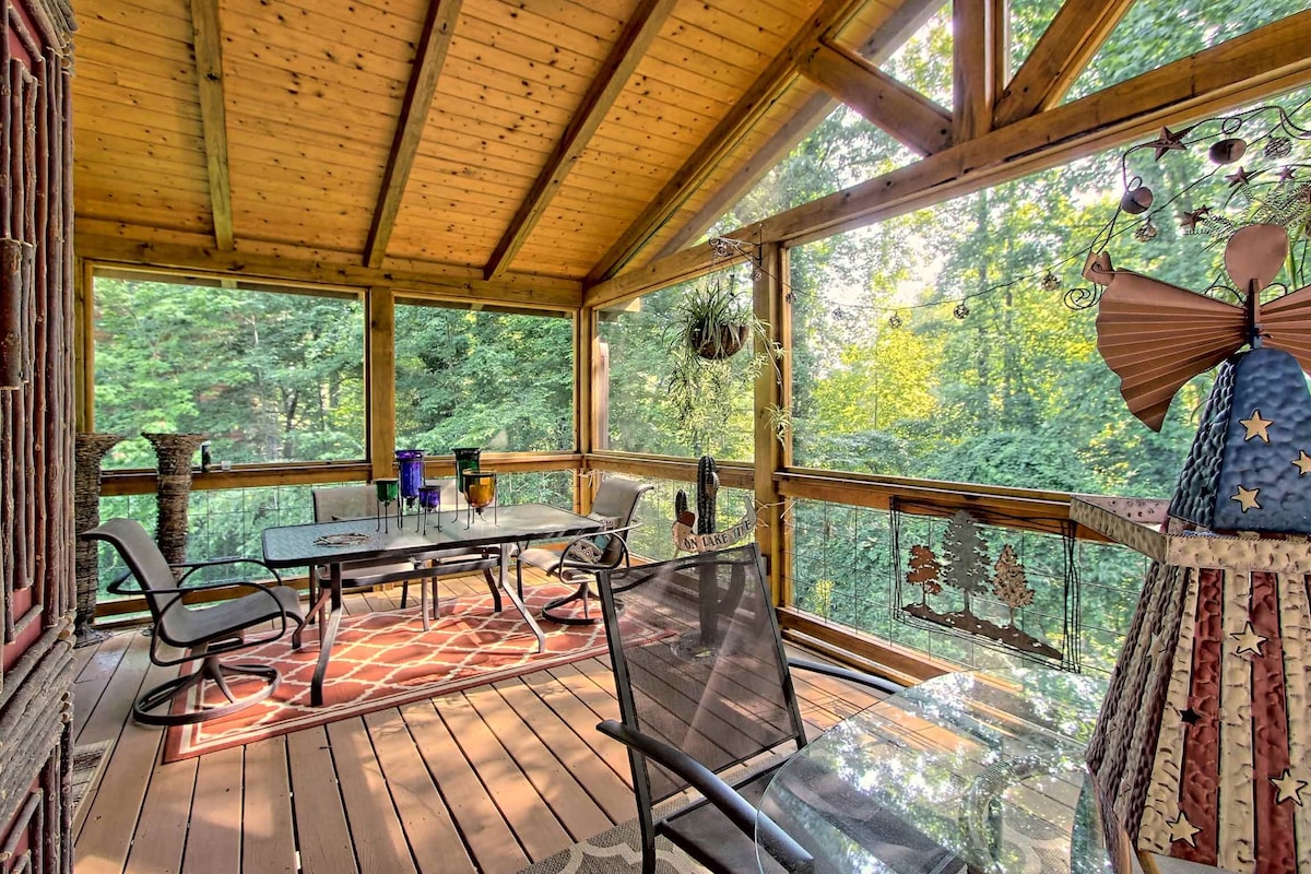 A screened porch features a wooden ceiling and glass table set with six chairs. Natural light filters through the foliage, creating a serene atmosphere. Plants adorn the space, adding a touch of greenery, while decorative pieces enhance the inviting design.