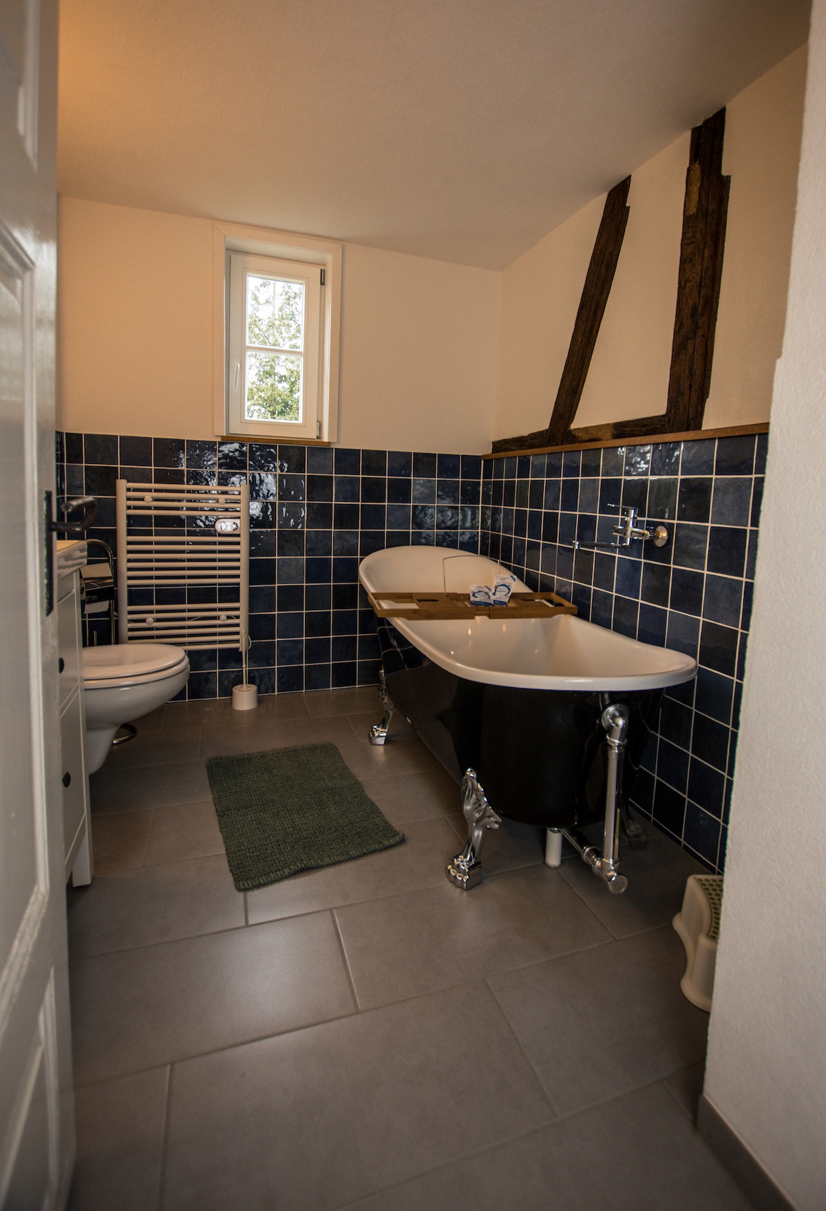 A modern bathroom features a freestanding bathtub with elegant chrome feet against a backdrop of blue tiles. A heated towel rack is positioned next to the toilet, and natural light enters through a small window. Soft grey tiles offer a clean, welcoming look.