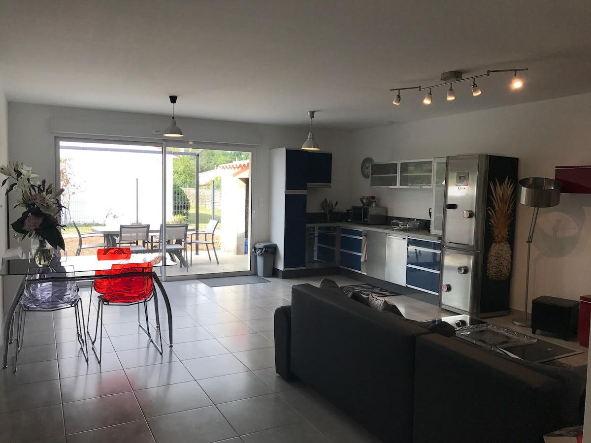 A modern living space is featured, showcasing a sleek kitchen with stainless steel appliances. A dining area is highlighted with transparent red chairs surrounding a glass table. Natural light enters through large windows, offering views of the outdoor terrace and garden.