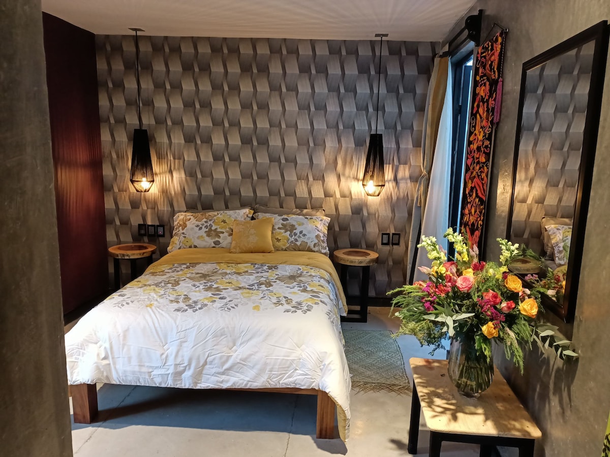 A cozy sleeping area features a floral-patterned bedding set on a spacious bed. Soft lighting is provided by pendant lamps on either side, while a mirror reflects a wooden side table. A vase of colorful flowers adds a touch of warmth to the room.