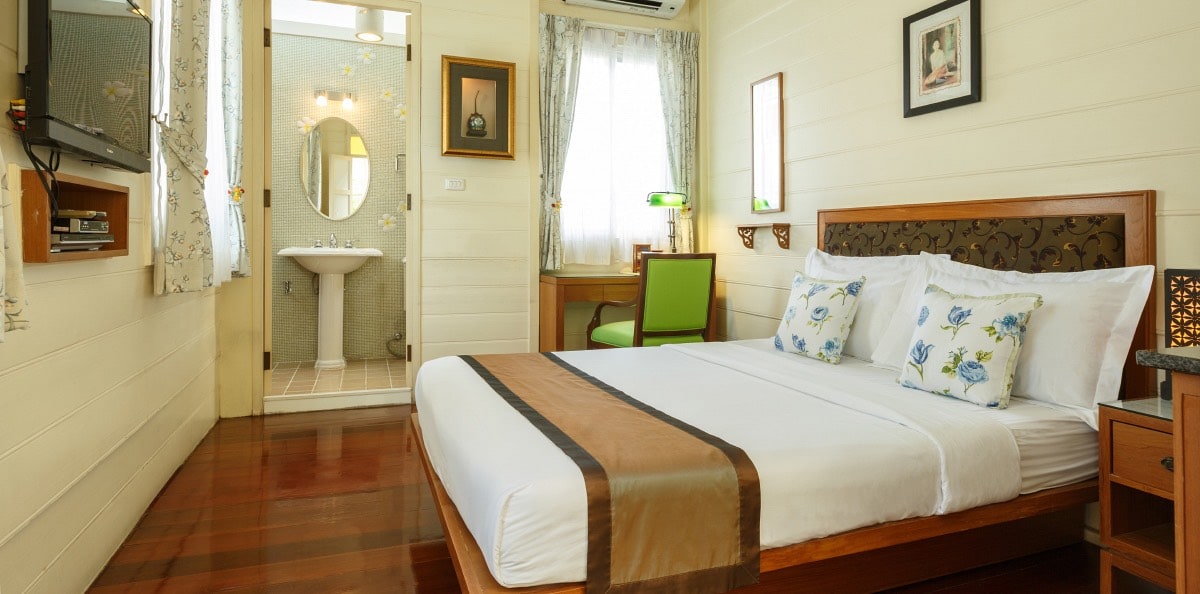 A spacious room features a queen size bed with decorative pillows, wood accents, and polished wooden floors. Natural light filters through the curtains, illuminating a work desk with a green chair. A private bathroom is visible in the background, showcasing modern amenities.
