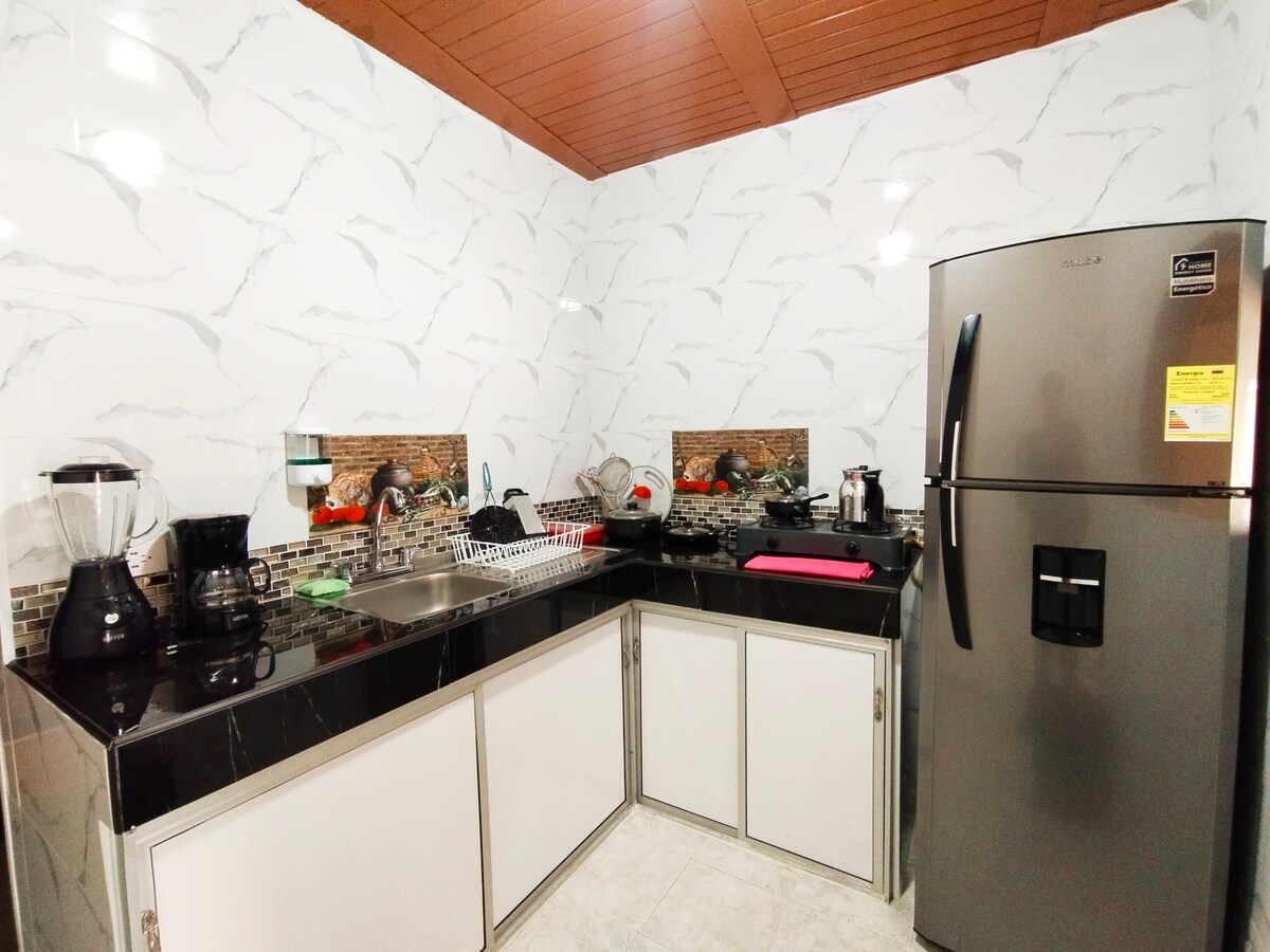 The kitchen is equipped with modern appliances, including a large refrigerator and a blender. Marble-patterned walls complement the sleek, dark countertops. A stainless steel sink is situated near a window, and various cooking utensils and dishes are neatly organized for convenience.