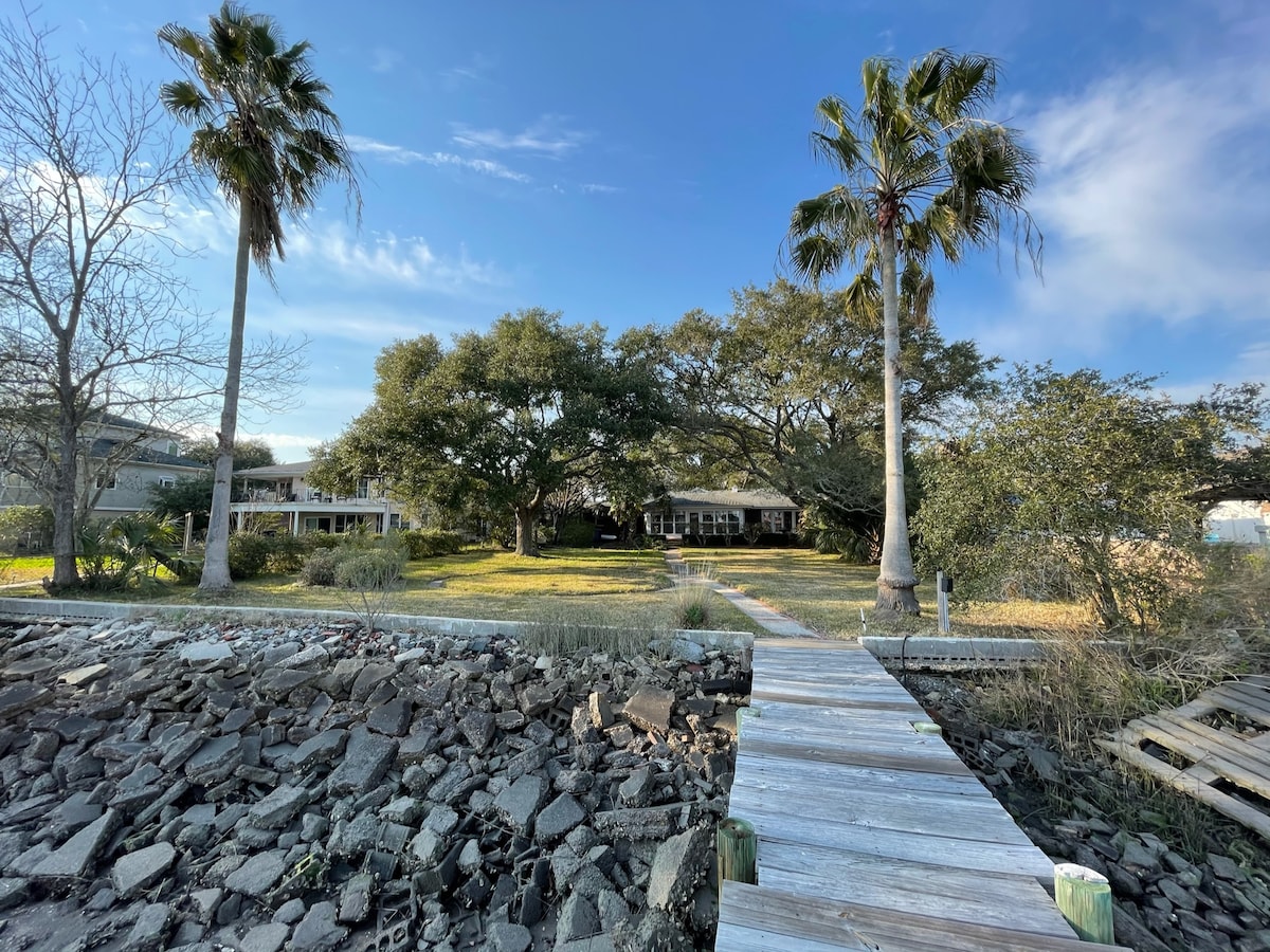 A wooden dock leads to a waterfront home surrounded by greenery, featuring large live oaks and palm trees. The landscape includes a stone pathway that guides visitors through the lush yard, highlighting the serene setting near the water.
