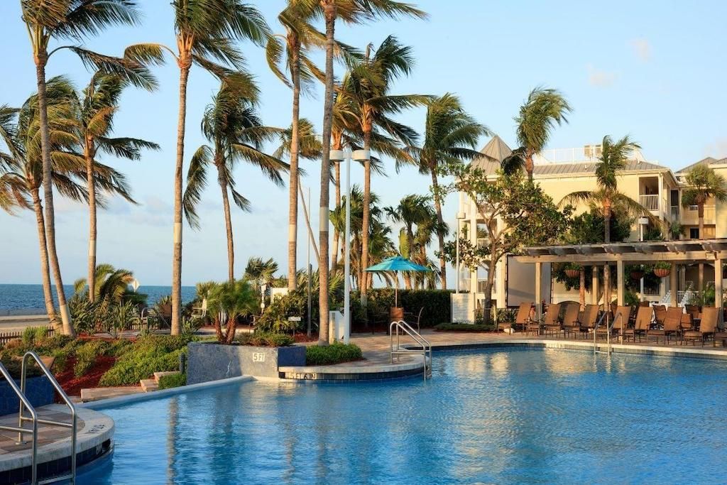 A serene pool area is surrounded by swaying palm trees, with inviting lounge chairs arranged for relaxation. The clear water reflects the sky, creating a tranquil atmosphere. A shaded seating area is visible nearby, adding to the vibrant landscape that leads towards the ocean.