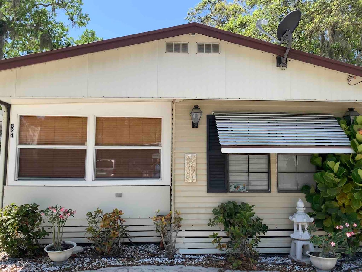 The exterior of a remodeled home features large front windows with wooden blinds and a welcoming entrance. Lush greenery and flowering plants are arranged along the pathway, while a decorative lantern and a small water feature add charm to the overall appeal.
