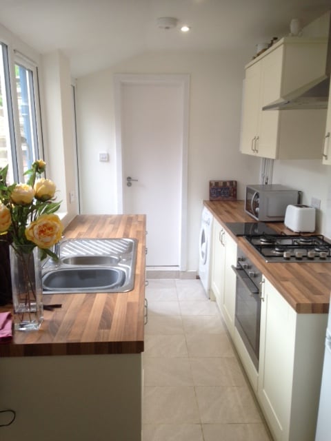 The kitchen features light-colored cabinetry and a wooden countertop. A double sink is positioned next to a modern stove and oven. Natural light streams in through the surrounding windows, while a washing machine is discreetly placed near the entrance door. Fresh flowers add a touch of elegance.