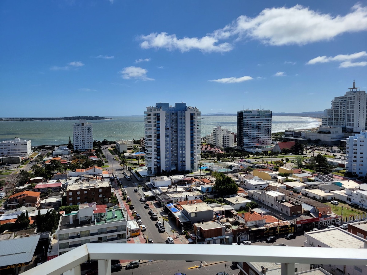 An expansive view of the coastline and cityscape is captured, showcasing a blend of residential and commercial buildings. The ocean reflects a clear blue sky with scattered clouds, while the streets below are lined with various structures, illustrating a vibrant urban setting.