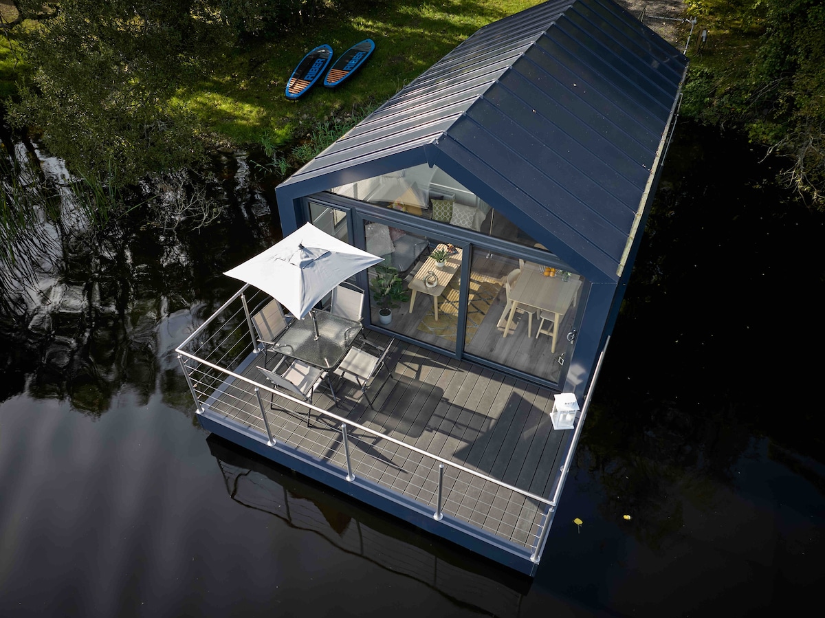 The houseboat is viewed from above, showcasing its modern design and outdoor balcony. A patio umbrella provides shade over a small seating area, and two kayaks rest nearby on the calm waters of Lough Erne, surrounded by lush greenery.