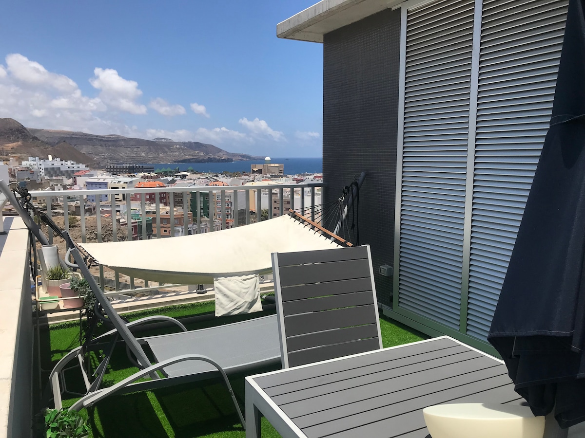A rooftop terrace is presented, featuring a hammock and lounge chairs. The view of the ocean and city skyline is visible in the background under a clear blue sky. The space is designed with artificial grass, providing a comfortable area to relax outdoors.