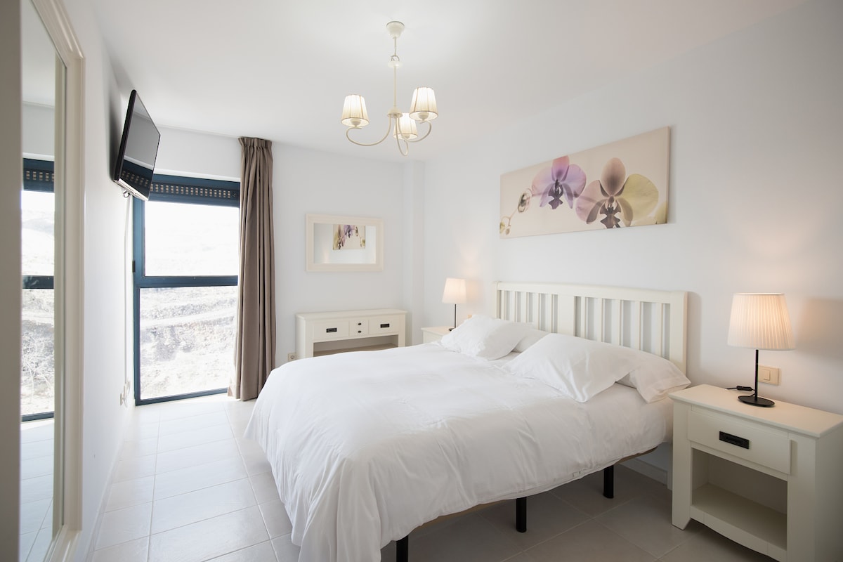 A bright and modern bedroom features a spacious bed with white bedding. A flat-screen TV is mounted on the wall, and soft lighting is provided by bedside lamps. Large windows offer natural light and a view of the outdoors, creating a calm atmosphere.