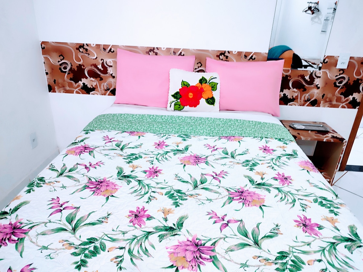 A comfortably furnished sleeping area features a floral-patterned bedspread and bright pink pillows against a decorative headboard. A small bedside table is visible, complementing the clean and organized space. A mirror reflects the room, enhancing the light and openness.