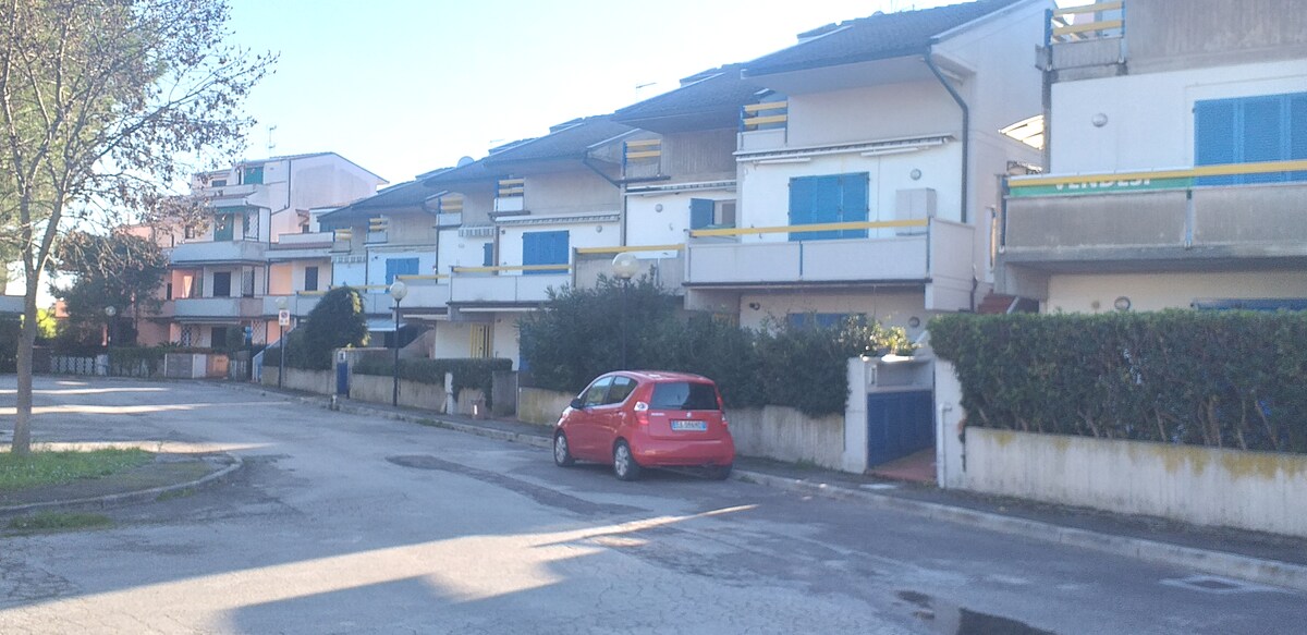 A row of modern multi-level buildings is displayed, featuring balconies with blue shutters. A red car is parked along the curving street, with greenery lining the pathway, providing a sense of neighborhood ambiance. Clear skies above create a bright, inviting atmosphere.