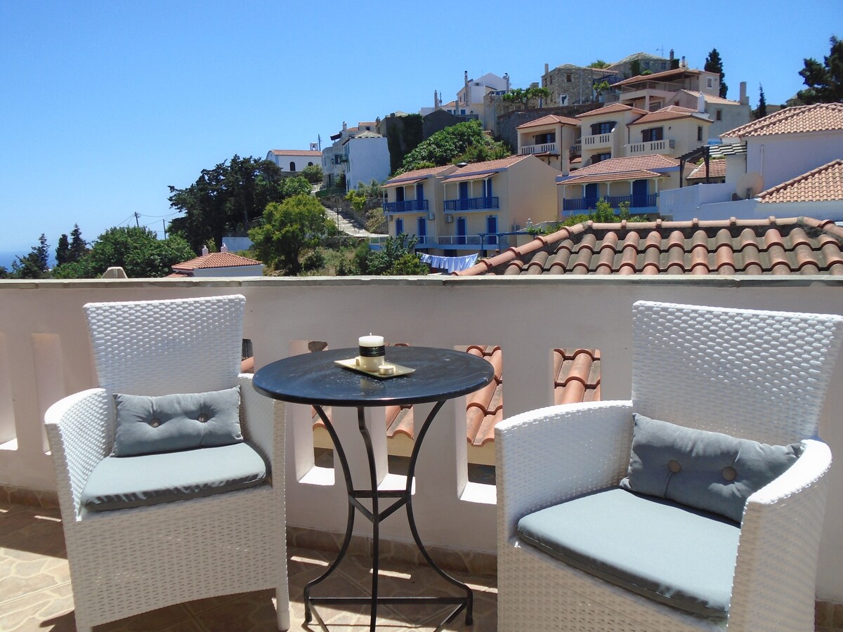 A spacious balcony is furnished with two white chairs and a small circular table, facing a scenic view of a traditional village and surrounding hills. Tile roofs and blue accents of nearby buildings are visible against a clear sky.