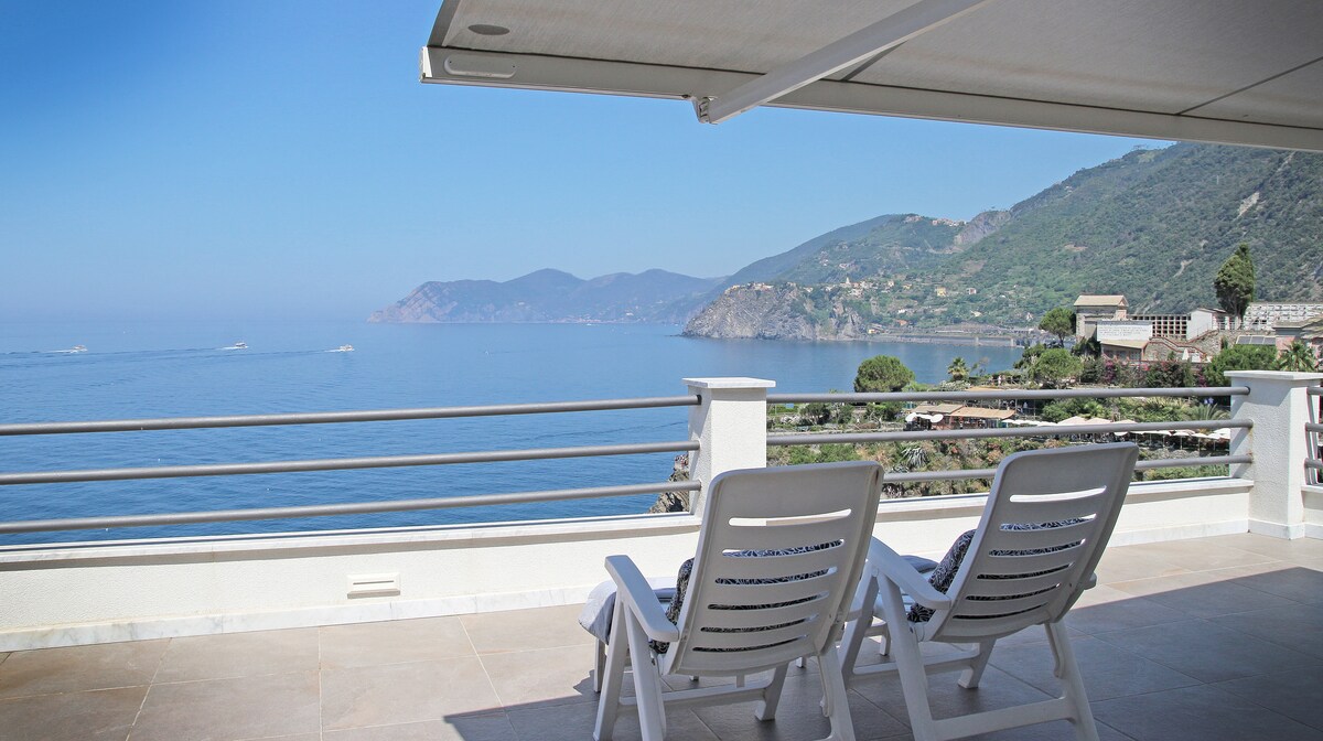 A terrace is shown with two loungers facing a panoramic view of the sea and the hills beyond. The clear blue water and distant coastline create a serene backdrop, while an awning provides shade over the seating area.
