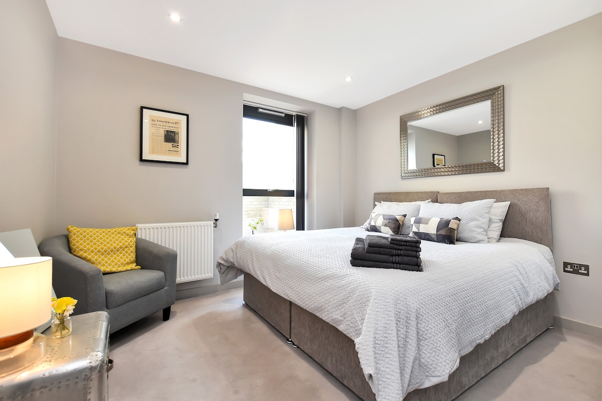 A spacious bedroom is displayed, featuring a large bed with top-of-the-line linens, accented by neatly arranged towels. A cozy armchair sits in one corner, accompanied by a small lamp. Natural light is introduced through the window, while soft tones of grey create a calm environment.