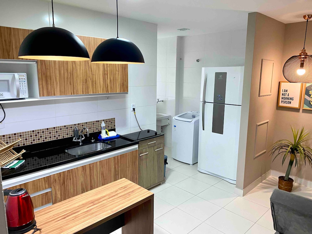 A modern kitchen area features wooden cabinetry paired with dark countertops. A stainless steel refrigerator and a washing machine are present, along with essential appliances like a microwave and kettle. Lighting fixtures hang above a wooden bar counter, enhancing the room's functionality.