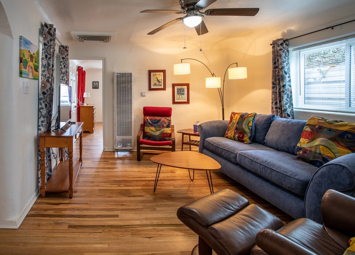 A cozy living area is presented with a plush blue sofa, complemented by patterned throw pillows and a wooden coffee table. A ceiling fan circulates air above, while warm lighting creates a comfortable ambiance. Nearby, a reclining chair and a standing air heater are visible.