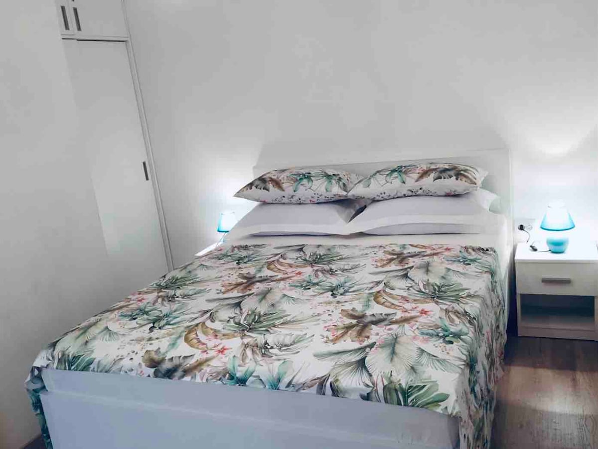 A bedroom features a spacious bed adorned with a floral patterned duvet and two matching pillows. Soft, ambient lighting is provided by two bedside lamps, enhancing the serene atmosphere. A closet is available for storage, contributing to the room's practical design.