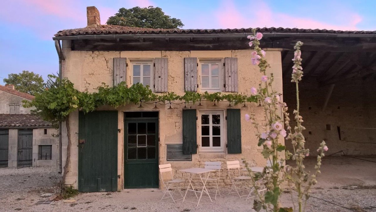 The rustic facade of an ecological gîte features a green door and window shutters, complemented by climbing plants. A gravel area in front supports a small, bistro-style table with two chairs, framing the entrance to the charming structure.