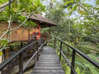 A wooden pathway leads toward a traditional Joglo house surrounded by lush greenery. Tropical plants frame the entrance, while a welcoming terrace with a thatched roof extends from the structure, providing a serene view of the vibrant garden.