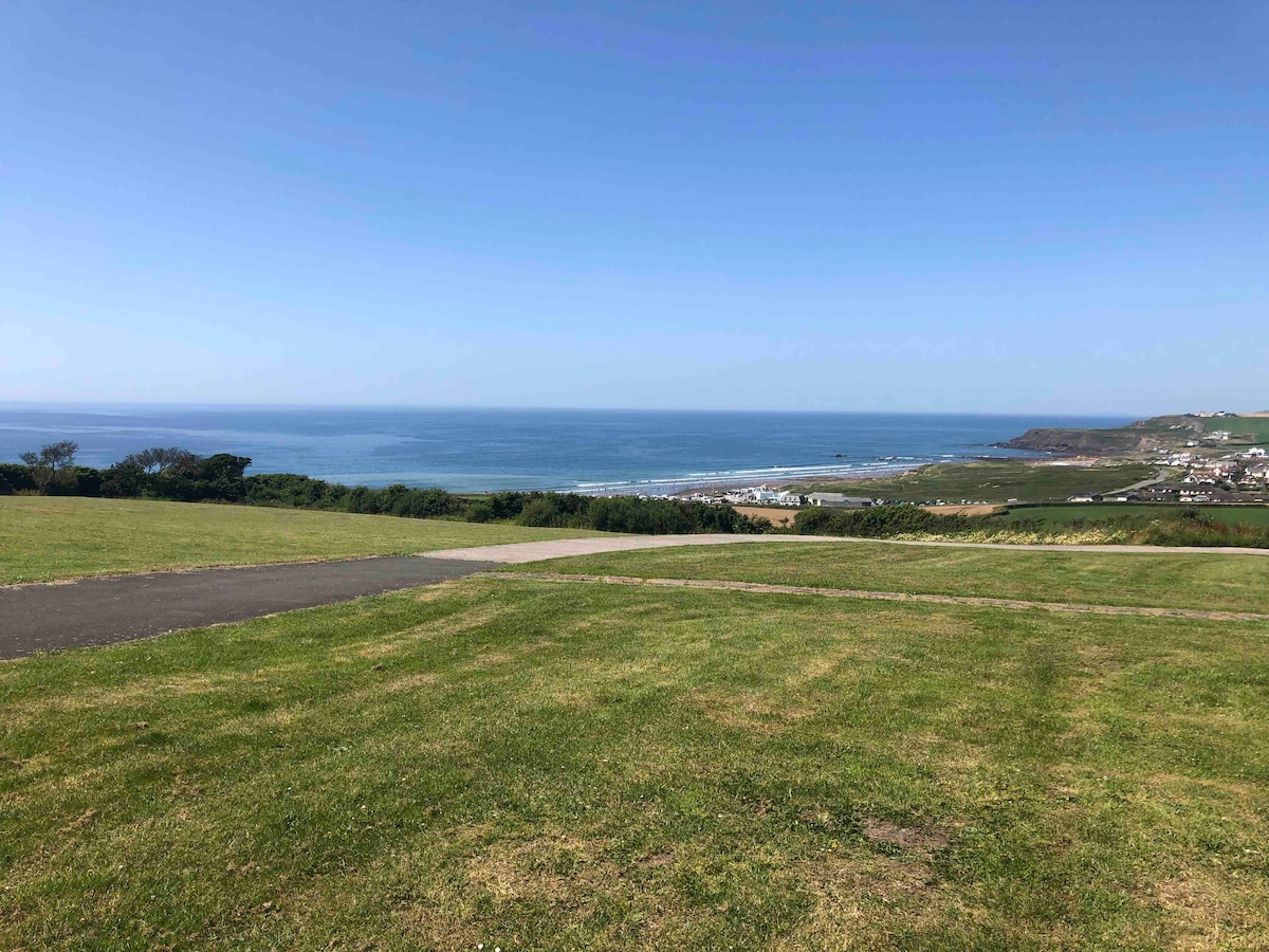 A panoramic view features the expansive sea stretching towards the horizon, with gentle waves visible. Lush green grass covers the foreground, leading to a paved pathway. The backdrop includes distant hills and coastal scenery, creating a serene coastal atmosphere.