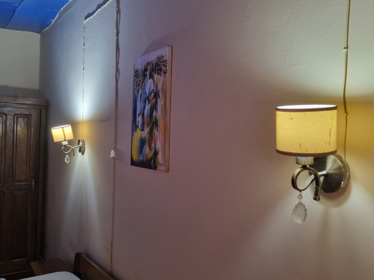 The image displays a cozy sleeping area featuring two wall-mounted lamps with fabric shades, providing soft illumination. An artwork is hung above the bed, adding a touch of color to the neutral walls. A wooden wardrobe is visible in the background.