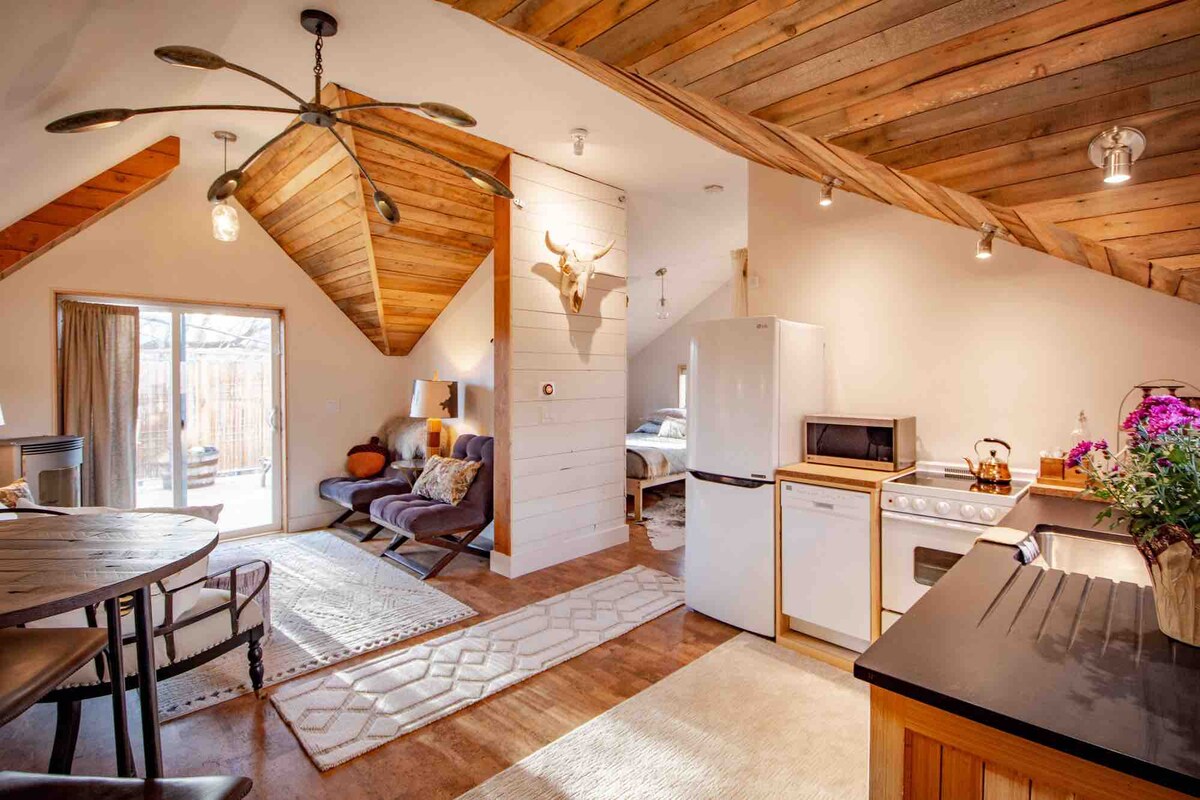 An open-concept living area features wood-beamed ceilings, a stylish kitchen with modern appliances, and a cozy seating arrangement. Natural light floods in from large glass doors leading to an outdoor space. A tasteful area rug anchors the space, enhancing its warm ambiance.