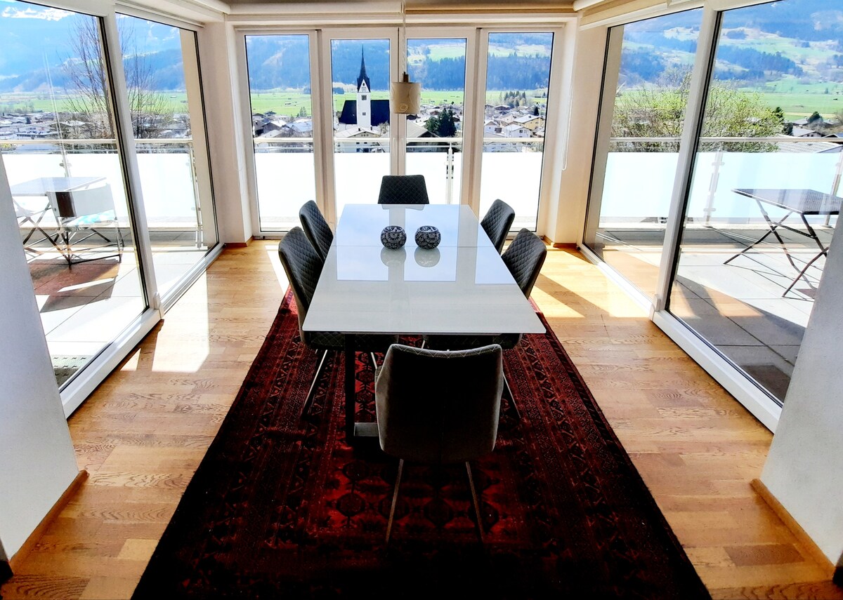 A dining area is illuminated by natural light, featuring a modern table surrounded by dark chairs. Floor-to-ceiling windows provide expansive views of the surrounding landscape, including a church steeple, while a Persian rug adds warmth to the wooden floor.