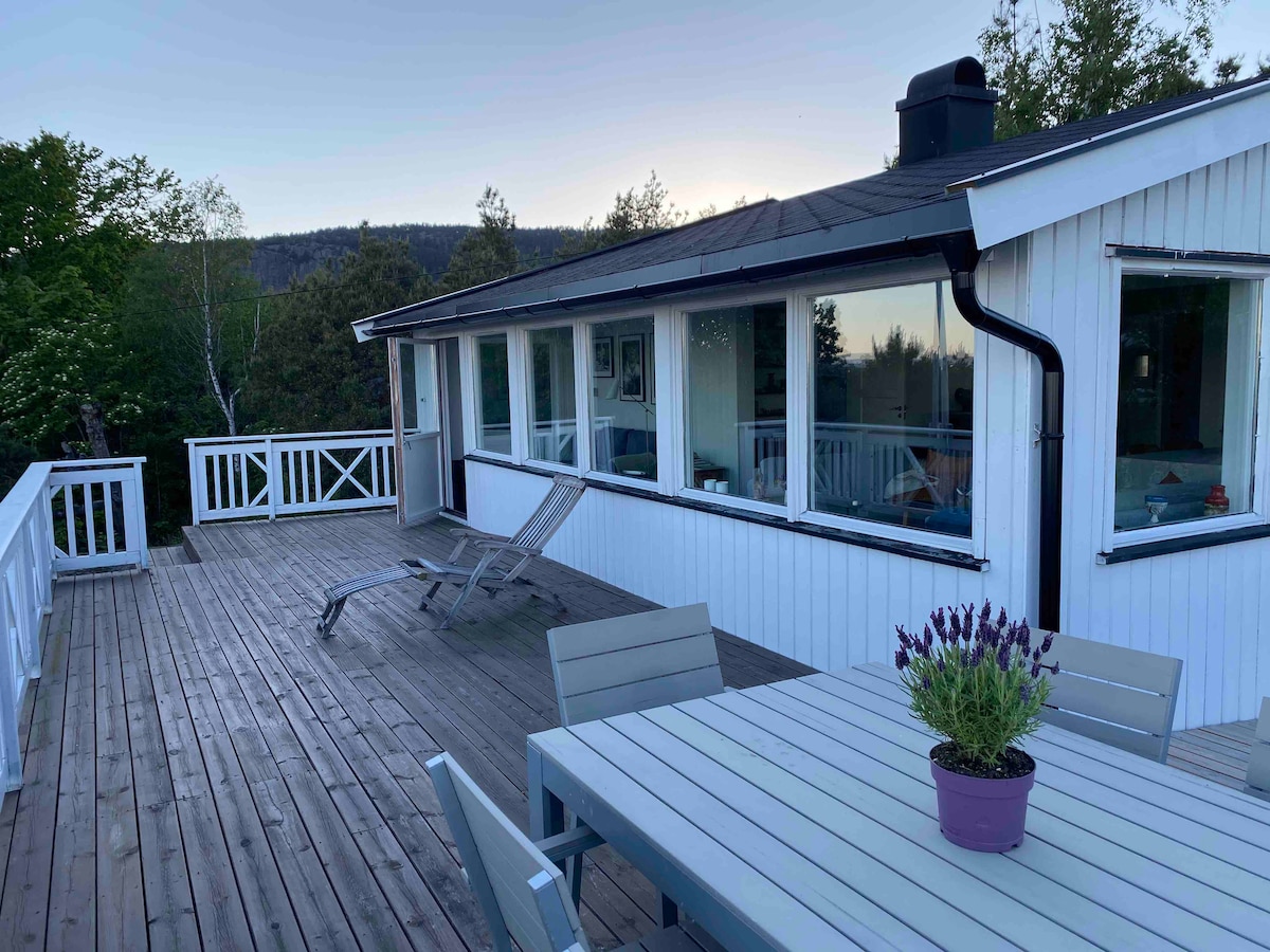 A spacious wooden terrace features a dining table with chairs and a lounge chair for relaxation. Large windows provide ample natural light, showcasing the interior of the cottage. Surrounding greenery enhances the tranquil outdoor space.