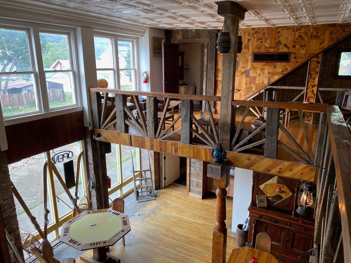 An open interior showcases a loft space with wooden railings above. Natural light filters in through large windows, illuminating the rustic decor, including wooden beams and a round table on the lower level. Various items are thoughtfully arranged to enhance the inviting atmosphere.